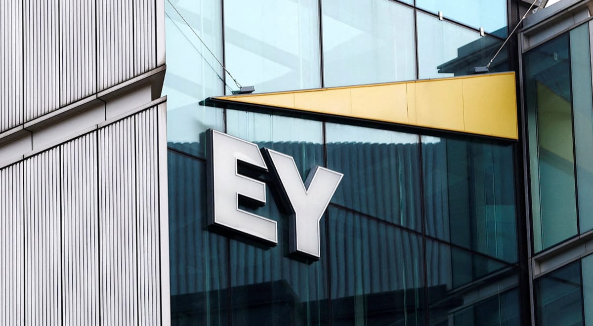 "EY" logo on the exterior of a modern glass building, reflecting the sky and surrounding architecture. The logo is prominent and appears to be a 3D white signage against the dark glass facade. A distinctive golden triangular architectural element is visible above the logo, adding to the building's modern aesthetic. This image is suitable for an article about the EY Internship 2025.